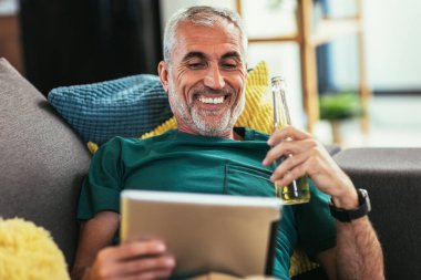 Olgun adam evdeki kanepede dinlenirken tablet bilgisayar kullanıyor.