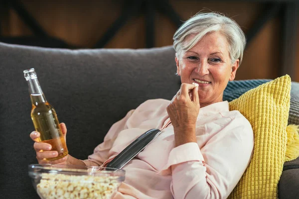 Senior woman enjoys watching tv at home. Senior woman watching tv drink beer and eat popcorn.