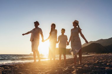 Aile kumsalda gün batımının tadını çıkarırken el ele tutuşuyor. Mutlu aile seyahatleri ve tatiller.