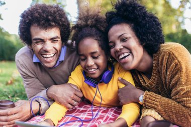 Güzel, genç bir aile piknik battaniyesinde uzanıp dijital tablet kullanırken sonbahar gününün tadını çıkarıyor..