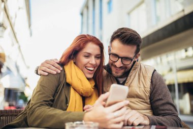 Happy young couple using a phone together at a coffee shop. Young man and woman looking at touch screen computer and smiling.
