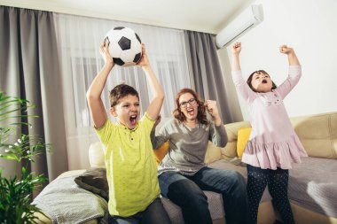 Mother and children with raising arms and screaming while watching football match and celebrating goal at home