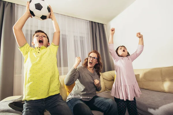 Mother and children with raising arms and screaming while watching football match and celebrating goal at home