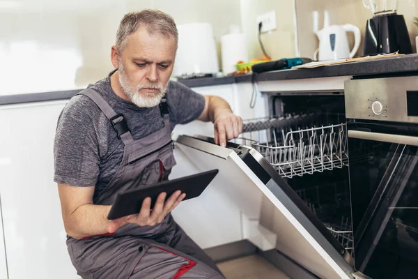 Portrait Of Male Technician Repairing Dishwasher In Kitchen using digital tablet.