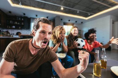 Young group watching sports on television and cheering. Emotional football fans watching football game together at home. Sports Fans with Painted Faces Sitting on a Couch Watch Game on TV