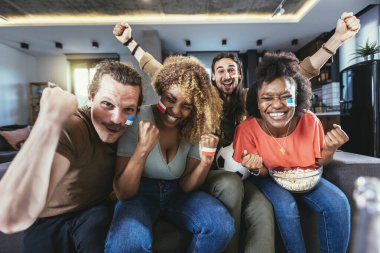 Young group watching sports on television and cheering. Emotional football fans watching football game together at home. Sports Fans with Painted Faces Sitting on a Couch Watch Game on TV