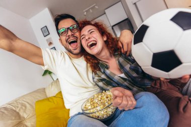 Mutlu genç çift hafta sonunu evde, kanepede oturup televizyonda futbol izleyerek ve favori takım için tezahürat yaparak geçiriyor.