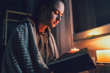 Genç kız elinde mumlarla kaloriferin yanında battaniyenin altında oturuyor ve kitap okuyor. Enflasyon ve savaş, enerji krizi ve enerji krizi nedeniyle doğal gaz faturası için özel evlerde artış maliyetleri