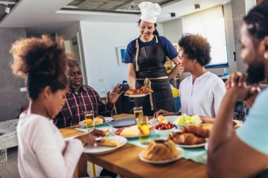 Yemek masasında yemek yiyen çok nesilli Afro-Amerikan bir aile. Beyaz kadın aşçı servis ediyor..