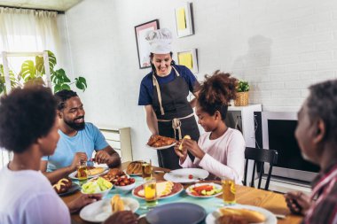 Yemek masasında yemek yiyen çok nesilli Afro-Amerikan bir aile. Beyaz kadın aşçı servis ediyor..