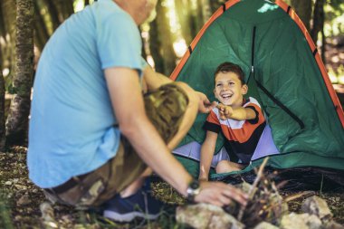 Baba ve oğlu dışarıda, orman ağaçlarının arka planında mutlu vakit geçiriyorlar. Birlikte kamp yapıyorlar ve eğleniyorlar.