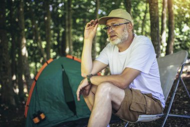 Arka planda bir çadırla ormanda oturan olgun bir adamın portresi. Olgun bir adam kamp alanında oturuyor..