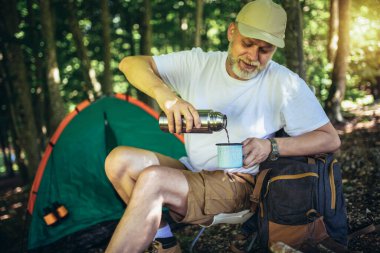 Ormanda arka planda çadırla oturan ve termostan kahve dolduran olgun bir adamın portresi. Olgun bir adam kamp alanında oturuyor..
