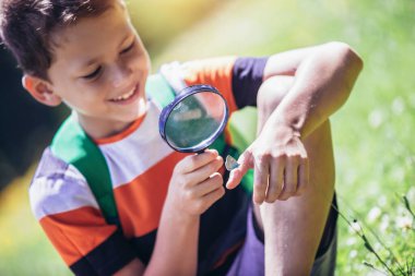 Genç bir çocuk büyüteçle çayırda doğayı keşfediyor.