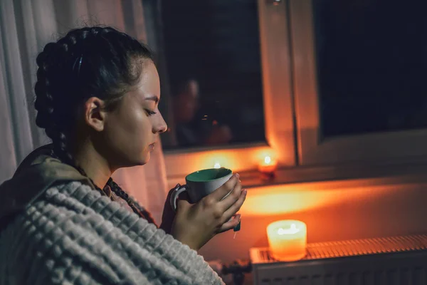 Teenage girl sits under blanket near heating radiator with cup of tea.Rising costs in private households for gas bill due to inflation and war, Energy crisis