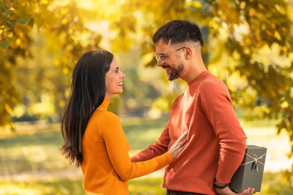 白人のボーイフレンドは 愛の愛情のバレンタインデーの誕生日のシンボルのために存在する秋の公園の驚きで美しいガールフレンドにボックスギフトを与えます — ストック写真