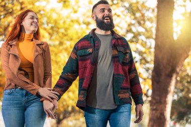Genç mutlu çift el ele tutuşup sonbahar parkında yürürken birbirlerine bakıyorlar..