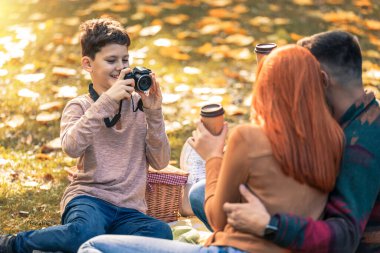 Genç mutlu ebeveynler sonbahar günü parkta oğluyla eğleniyorlar. Çocuk anne ve babasının piknikte fotoğrafını çekiyor. 