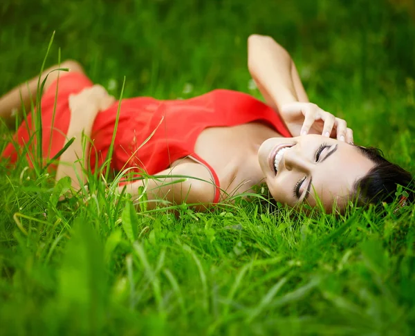 Mode porträtt av vacker sexig kvinnlig brunett tjej modell liggande i gröna sommaren ljusa gräs i parken med smink i röd dress.perfect skin.nature — Stockfoto