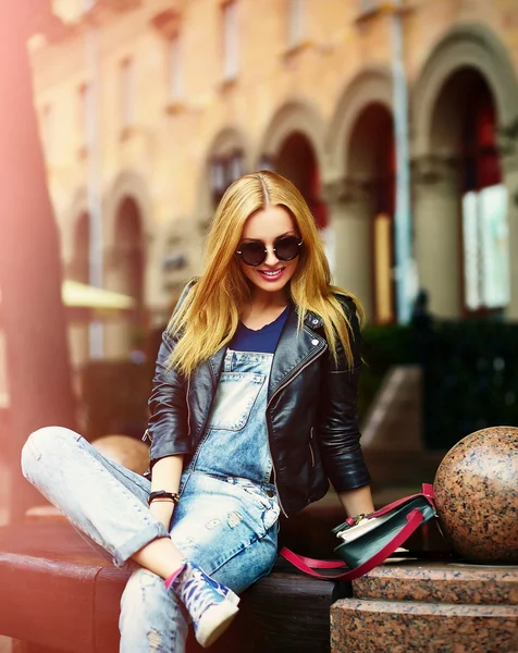 Retrato de bonito engraçado moderno sexy urbano jovem elegante sorridente mulher menina modelo em brilhante pano moderno ao ar livre sentado no parque em jeans em um banco de óculos — Fotografia de Stock