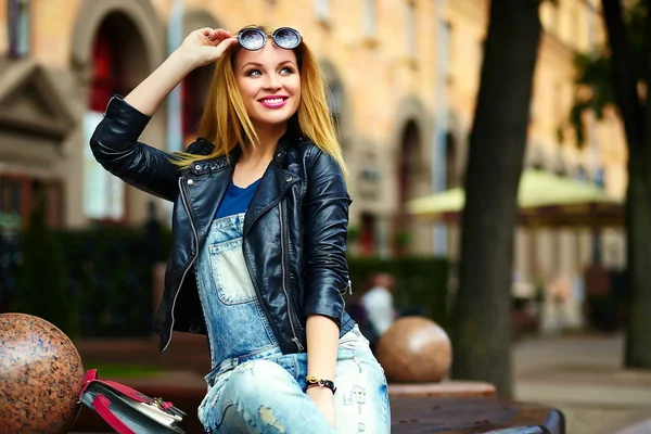 Retrato de lindo divertido moderno sexy urbano joven elegante mujer sonriente modelo de niña en tela moderna brillante al aire libre sentado en el parque en jeans en un banco en gafas — Foto de Stock