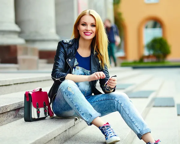 Retrato de lindo divertido moderno sexy urbano joven elegante mujer sonriente modelo de niña en tela moderna brillante al aire libre sentado en el parque en jeans en un banco en gafas con bolsa de color rosa —  Fotos de Stock