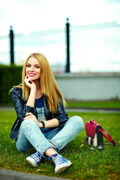 Retrato de lindo divertido rubio moderno sexy urbano joven elegante mujer sonriente modelo de niña en tela moderna brillante al aire libre sentado en el parque en jeans con bolsa de color rosa — Foto de Stock