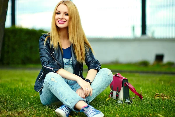 Retrato de lindo divertido rubio moderno sexy urbano joven elegante mujer sonriente modelo de niña en tela moderna brillante al aire libre sentado en el parque en jeans con bolsa de color rosa — Foto de Stock