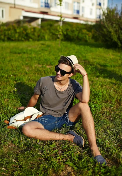 画像 》 吸引现代时尚的年轻人在休闲布在眼镜坐在公园里的绿草的帽子 — 图库照片