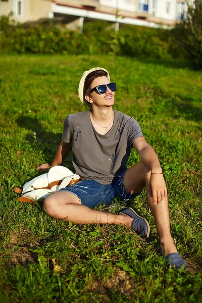Portrait of young attractive modern stylish man in casual cloth in hat in glasses sitting in the park in green grass — Stock Photo, Image