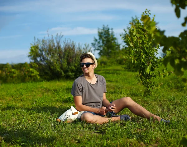 Portrait of young attractive modern stylish man in casual cloth in hat in glasses sitting in the park in green grass — Stock Photo, Image