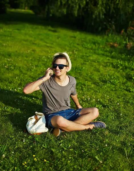 Портрет молодого привлекательного современного стильного мужчины в повседневной одежде в шляпе в очках, сидящего в парке в зеленой траве — стоковое фото