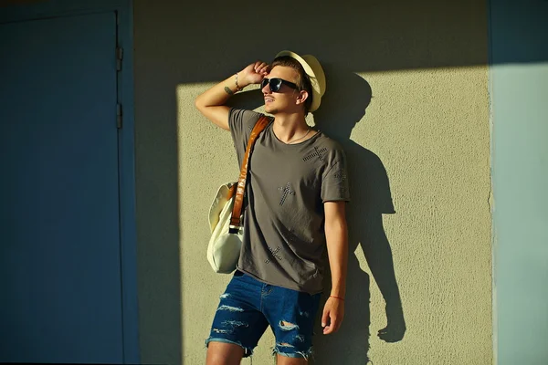 Retrato de jovem atraente sorrindo moderno elegante homem em pano casual em chapéu em óculos de sol de pé perto da parede — Fotografia de Stock