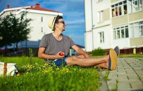 Casual kumaş şapkalı yeşil çimen parkta oturan güneş gözlüklü genç çekici modern şık adam portresi — Stok fotoğraf