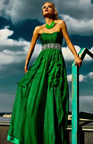 Aspecto de alta moda. sexy modelo de mujer joven rubia elegante con labios rojos maquillaje brillante con perfecta piel bañada por el sol con joyas al aire libre en estilo de moda en vestido largo verde de noche detrás del cielo —  Fotos de Stock