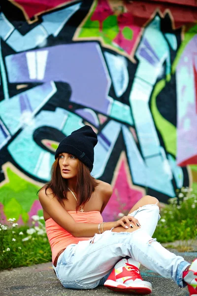 Retrato de sexy urbano moderno joven mujer elegante modelo de niña en tela moderna brillante en jeans al aire libre en la calle detrás del graffito —  Fotos de Stock