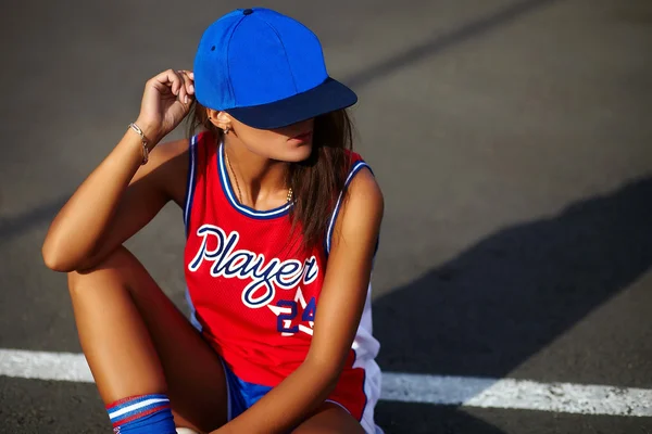 Portrait de sexy urbain moderne jeune femme élégante fille modèle en tissu sport de basket-ball rouge décontracté lumineux à l'extérieur assis dans la rue en bonnet bleu — Photo