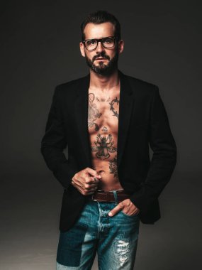 Yakışıklı, kendine güvenen, stil sahibi hipster Lambersex modelinin portresi. Siyah ceket giyen seksi modern adam. Dövmeli çıplak gövde. Stüdyoda siyah arka planda poz veren moda erkek. Gözlüklerle