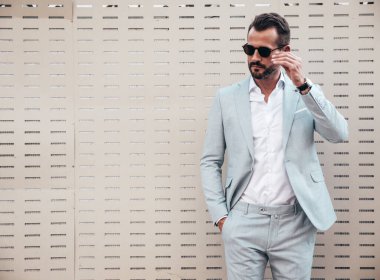 Yakışıklı, kendine güvenen, stil sahibi, lamberseksüel modelin portresi. Zarif beyaz takım elbise giymiş modern bir adam. Moda erkeği gün batımında çitlerin yanında poz veriyor. Güneş gözlükleriyle