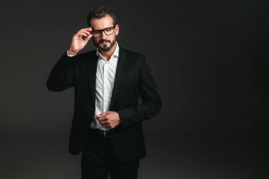 Yakışıklı, kendine güvenen, stil sahibi hipster Lambersex modelinin portresi. Zarif siyah takım elbiseli seksi modern adam. Moda erkeği karanlık arka planda stüdyoda poz veriyor. Gözlüklerle