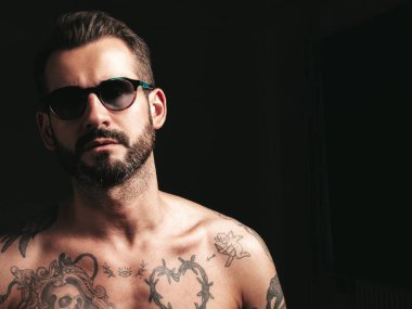 Yakışıklı, kendine güvenen, stil sahibi hipster Lambersex modelinin portresi. Seksi, modern adam. Dövmeli çıplak gövde. Stüdyoda siyah arka planda poz veren moda erkek. Güneş gözlükleriyle