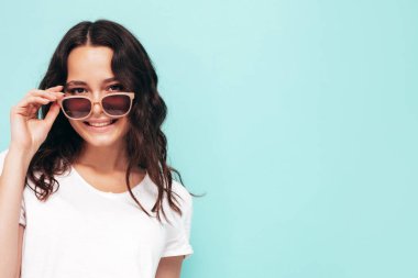 Moda bir yaz elbisesi içinde gülümseyen genç bir kadının yakın plan portresi. Stüdyoda mavi duvarın yanında poz veren kaygısız bir kadın. Kapalı alanda eğlenen pozitif bir model. Neşeli ve mutlu. Güneş gözlüğünü çıkarır.