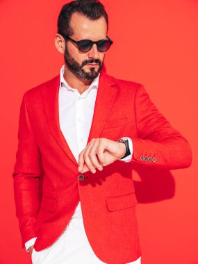 Yakışıklı, kendine güvenen, stil sahibi hipster Lambersex modelinin portresi. Zarif takım elbiseli seksi modern adam. Stüdyoda güneş gözlüklü, kırmızı duvarın yanında poz veren moda erkek. Saatlere bakıyorum. 