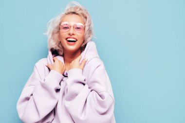 Son moda yaz kapüşonlu güzel, gülümseyen bir kadın. Stüdyoda mavi duvarın yanında poz veren kaygısız seksi kadın. Pozitif sarışın model eğleniyor ve deliriyor. Neşeli ve mutlu. Güneş gözlükleriyle