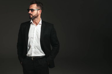 Yakışıklı, kendine güvenen, havalı hipster Lambersex modelinin yakın plan portresi. Zarif siyah takım elbiseli seksi modern adam. Moda erkeği karanlık arka planda stüdyoda poz veriyor. Güneş gözlükleriyle