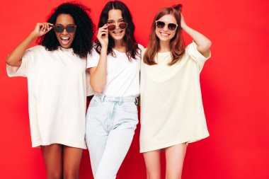 Moda kıyafetleri içinde gülümseyen üç güzel uluslararası hippi kadın. Stüdyoda kırmızı duvarın yanında poz veren kaygısız seksi kadınlar. Pozitif modeller eğleniyor. Arkadaşlık kavramı