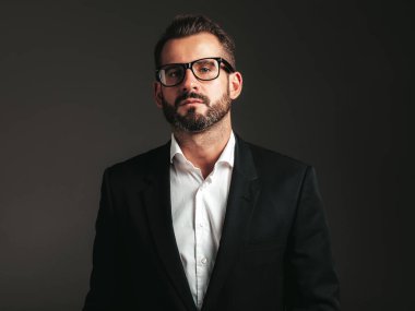 Yakışıklı, kendine güvenen, stil sahibi hipster Lambersex modelinin portresi. Zarif siyah takım elbiseli seksi modern adam. Moda erkeği karanlık arka planda stüdyoda poz veriyor. Gözlüklerle
