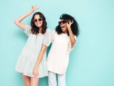 Moda kıyafetleri içinde iki güzel, gülümseyen uluslararası hippi kadın. Stüdyoda mavi duvarın yanında poz veren kaygısız seksi kadınlar. Pozitif modeller eğleniyor. Arkadaşlık kavramı. Güneş gözlükleriyle