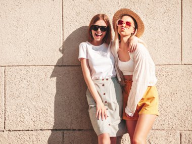 Son moda yaz kıyafetleri içinde iki güzel, gülümseyen hippi kadın. Sokakta beyaz duvarın yanında poz veren kaygısız kadınlar. Pozitif saf modeller gün batımında eğleniyorlar, sarılıyorlar ve deliriyorlar.