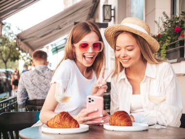 Son moda yaz kıyafetleri içinde iki güzel, gülümseyen yeniyetme kadın. Sokakta Veranda Kafe 'de oturan kaygısız kadınlar. Beyaz şarap içen, kruvasan yiyen, telefon ekranına bakan pozitif modeller.